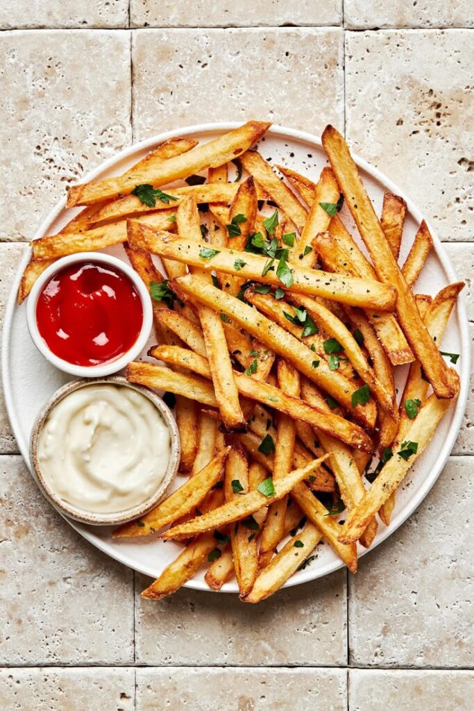 Crispy golden fries with parsley garnish served with ketchup and creamy dip on a white plate on stone tile background.