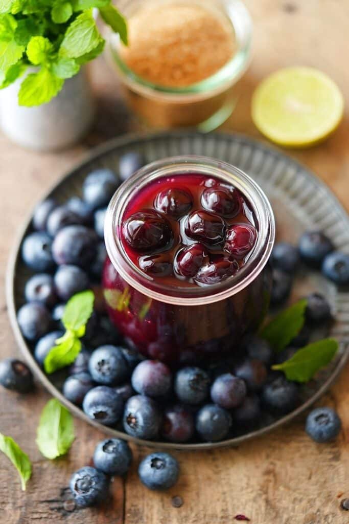 Jar of homemade blueberry jam surrounded by fresh blueberries and mint leaves on a rustic wooden table.