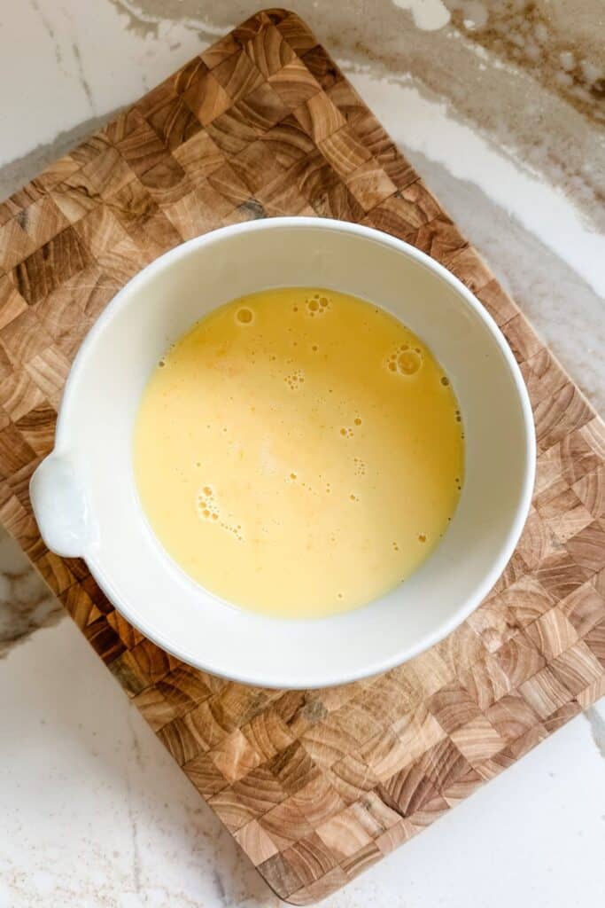 Beaten eggs in a white bowl on a wooden board, perfect for scrambled eggs or baking recipes.