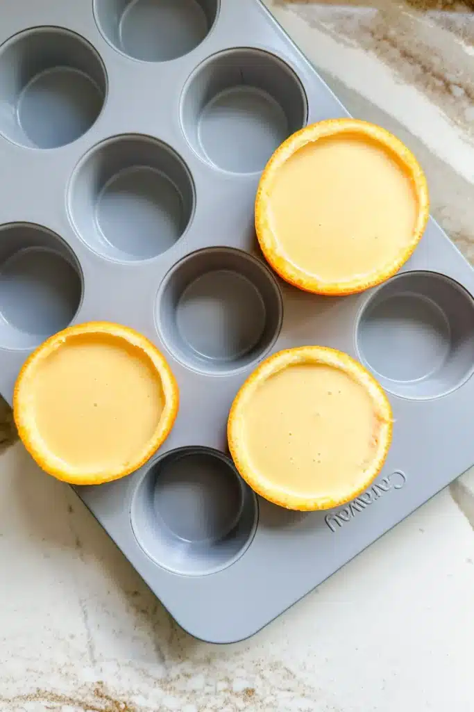 Orange slices filled with batter in a muffin tin on a marble countertop, ready for baking.