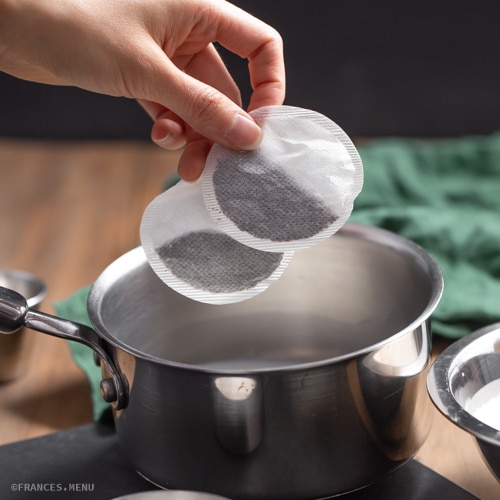 Hand holding tea bags above a saucepan on a wooden table, ready for brewing.