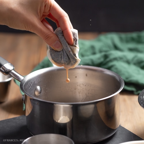 Hand squeezing a food pouch over a saucepan on a kitchen counter, preparing a culinary dish.