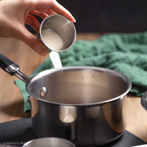 Hand adding ingredient into saucepan on stove, green cloth in background.