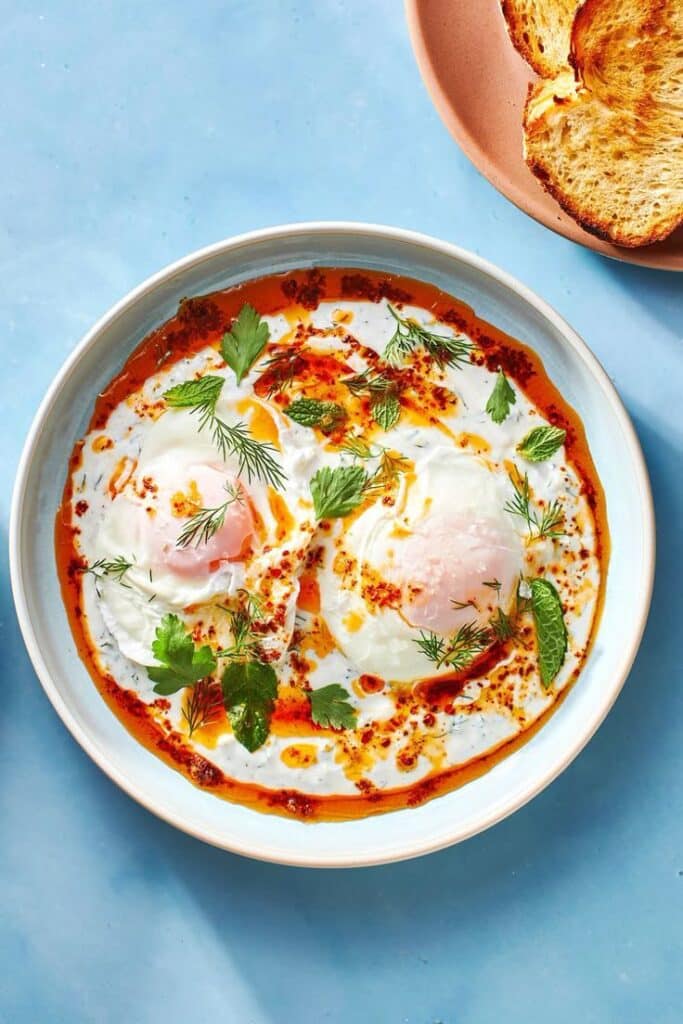 Delicious poached eggs in spicy yogurt sauce, garnished with herbs on a blue background with toast nearby.