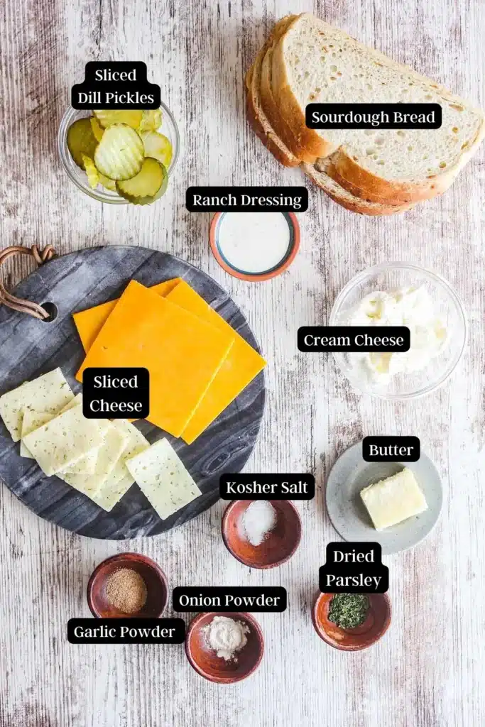 Ingredients for a sandwich: sourdough, cheese, pickles, cream cheese, ranch, butter, spices on a rustic surface.