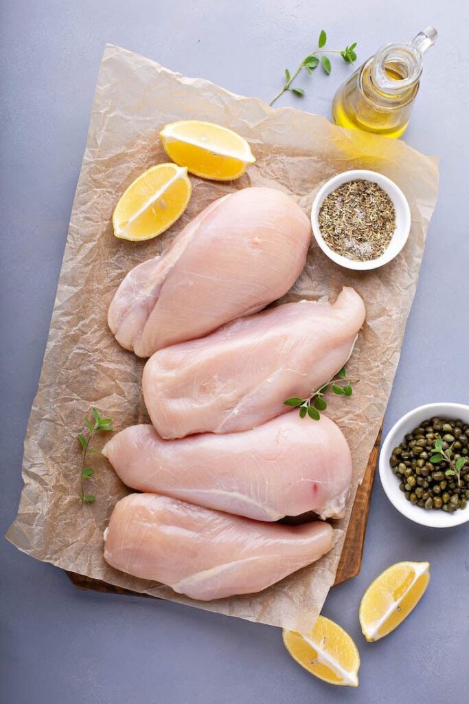 Raw chicken breasts on parchment with lemon, herbs, olive oil, and spices, ready for cooking preparation.