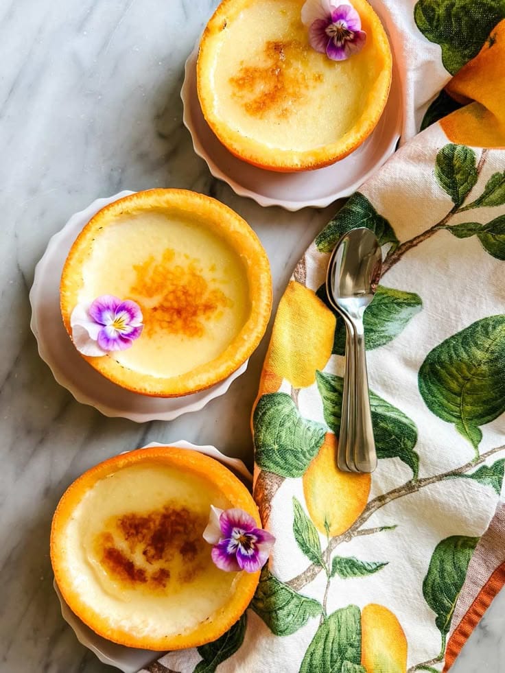 Creamy custard desserts in orange halves with edible flowers, on a floral napkin with a spoon, on a marble surface.