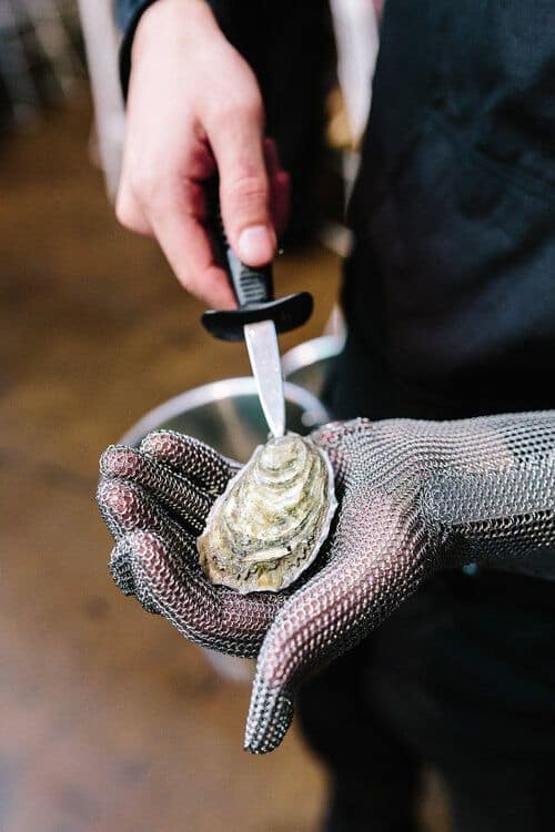 Person shucking an oyster with a knife, wearing a protective mesh glove for safety.