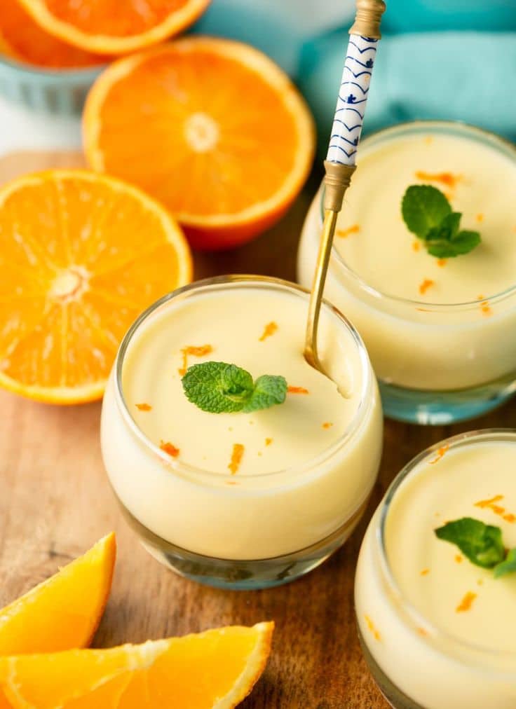 Creamy orange mousse in glass jars, garnished with mint leaves and fresh orange slices on a wooden table.