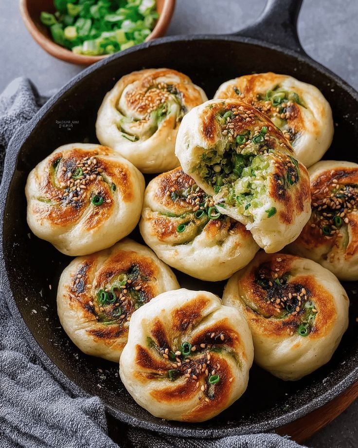 Pan-fried buns in a skillet, topped with sesame seeds and green onions, with one bun revealing a savory filling.