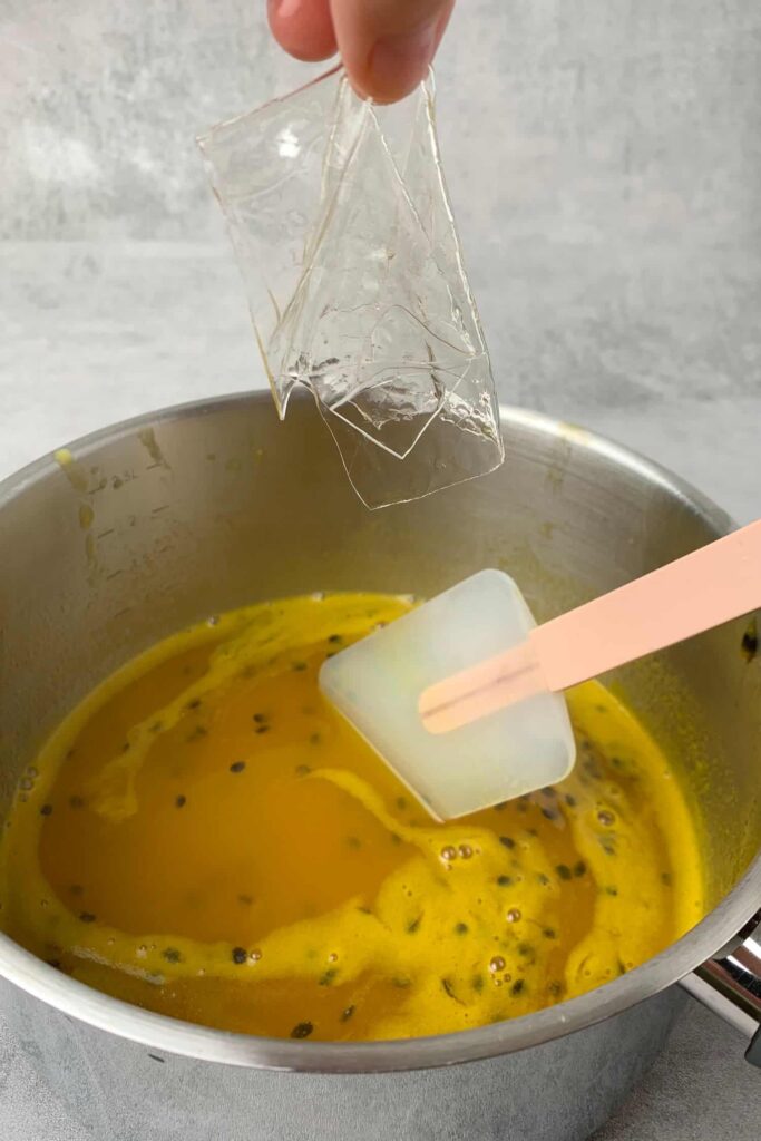 Passion-fruit-panna-cotta_2 - Silky, Tropical Coconut & Passion Fruit Panna Cotta Hand adding gelatin sheets to passion fruit mixture in a saucepan with spatula.