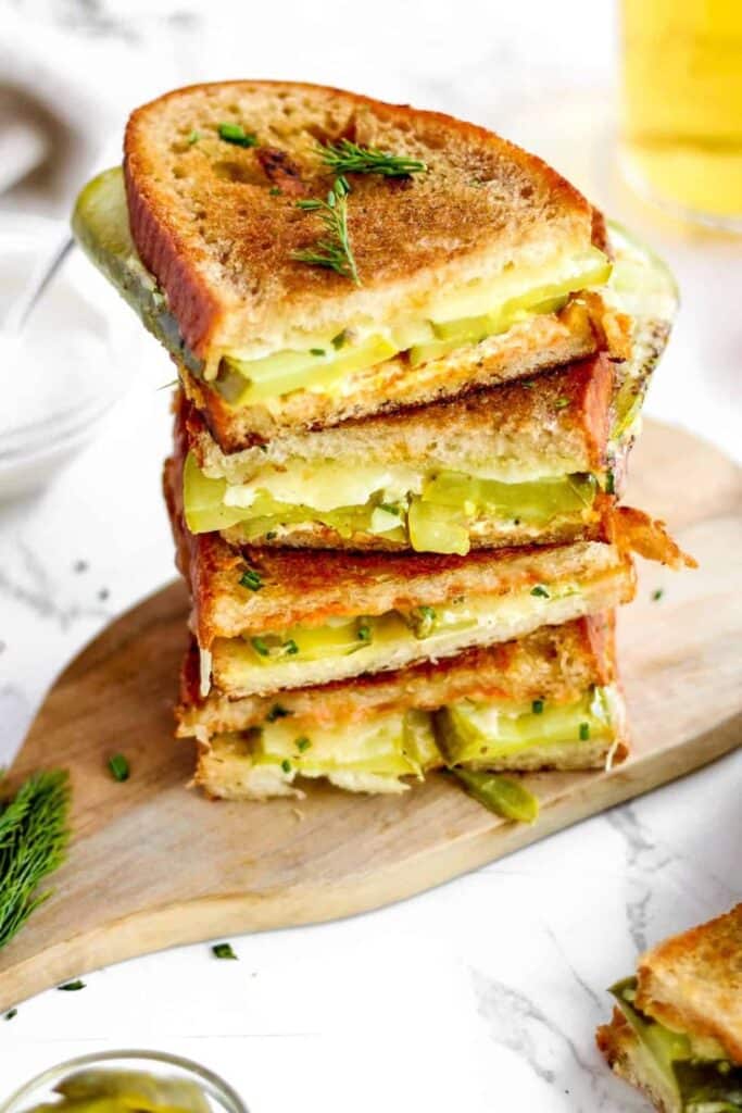 Stacked grilled cheese sandwiches with pickles on a wooden board, garnished with dill.