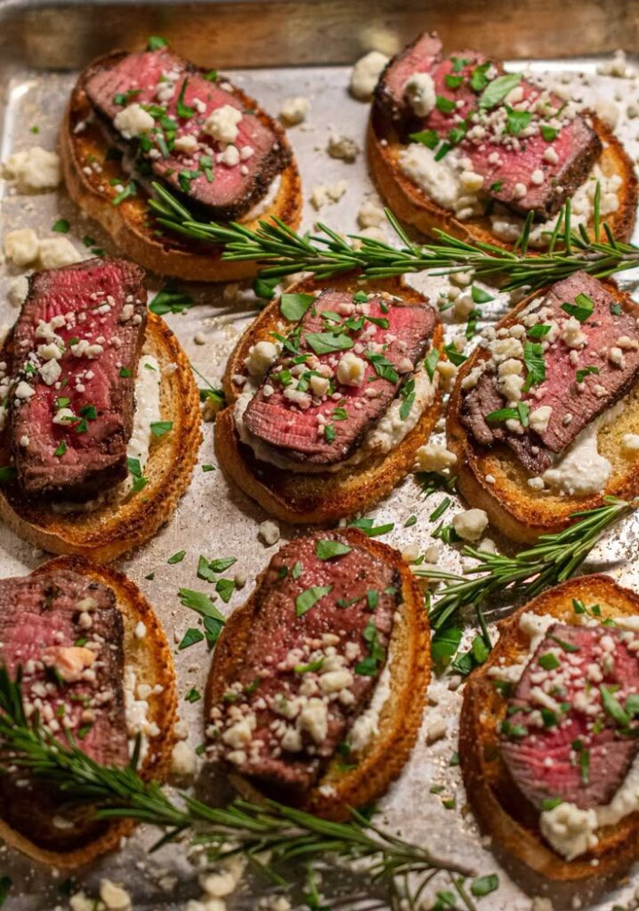 Beef crostini with cheese and herbs on a baking tray, garnished with rosemary and parsley. Appetizer recipe idea.