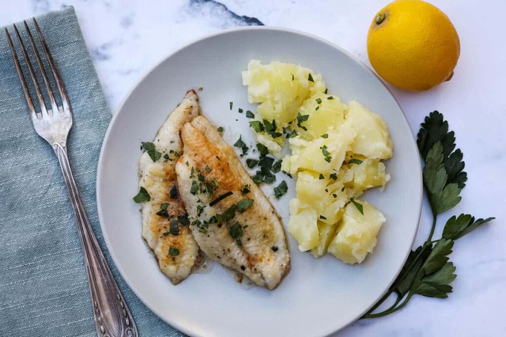 Sole-Meuniere-NEW2-1 - Enjoy a French Favorite: Sole Meunière & Potatoes Grilled fish fillets with herbs and mashed potatoes on a plate, garnished with parsley and a lemon on the side.