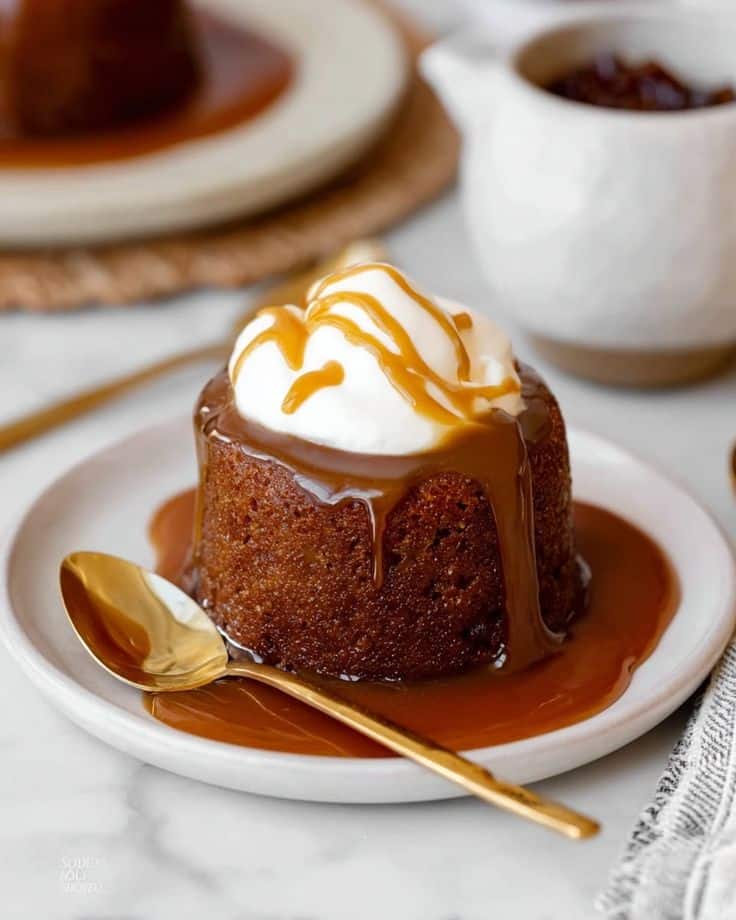 Sticky toffee pudding topped with whipped cream and caramel sauce, served on a white plate with a gold spoon.