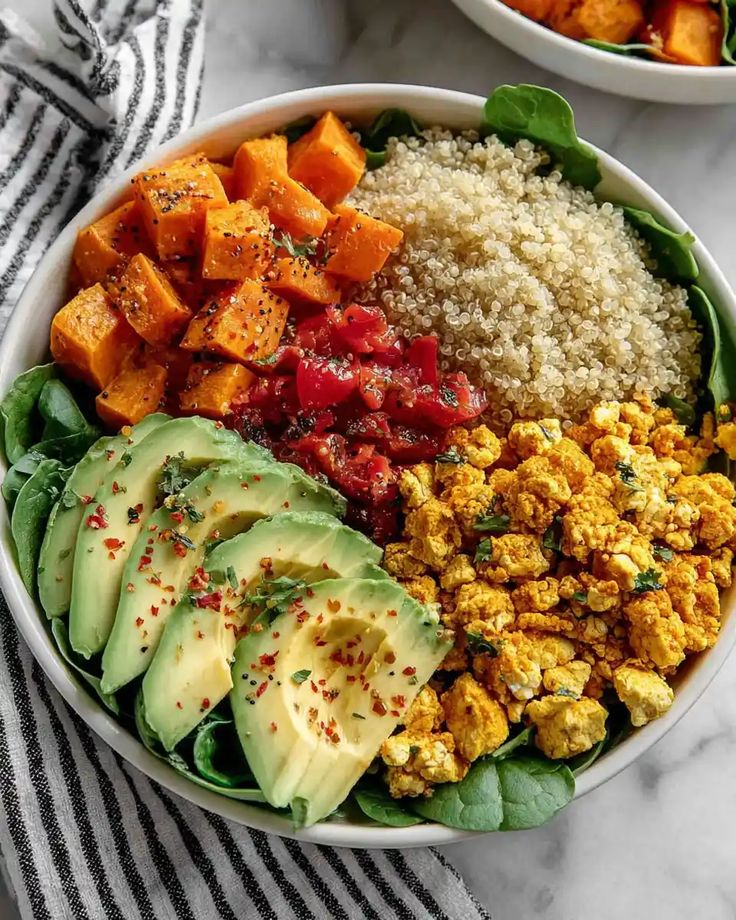 Vibrant vegan Buddha bowl with avocado, quinoa, sweet potatoes, roasted vegetables, and tofu scramble on a striped cloth.