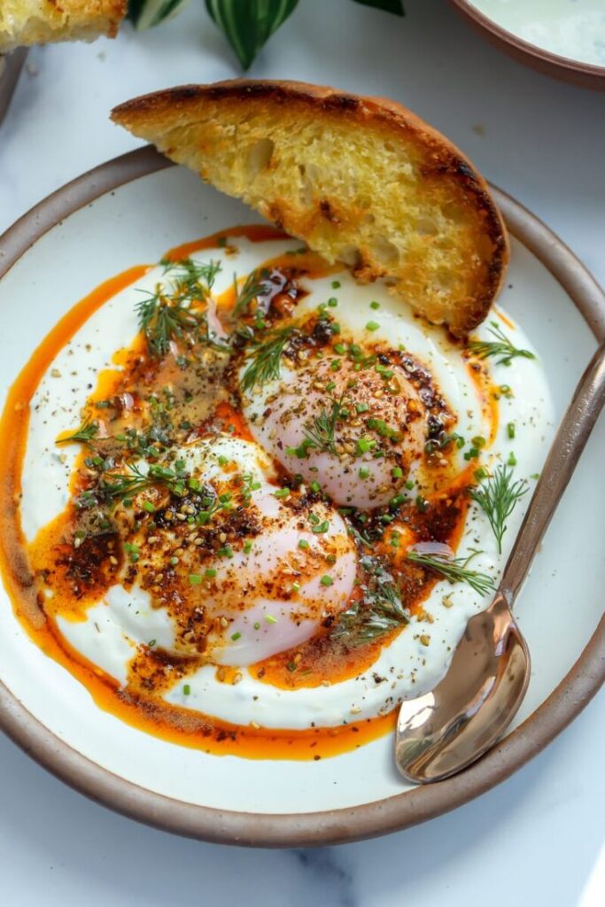 Poached eggs on yogurt with spices and herbs, served with toasted bread and garnished with fresh dill on a plate.