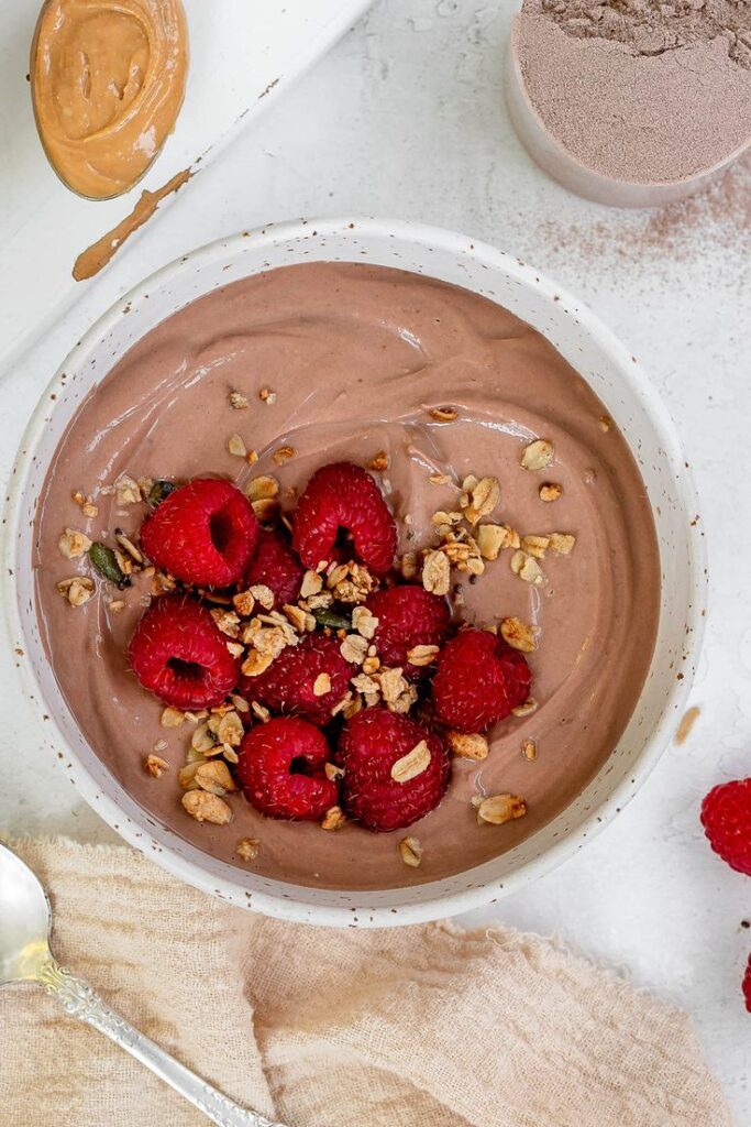 Chocolate protein smoothie bowl with raspberries and granola, surrounded by ingredients on a white surface.