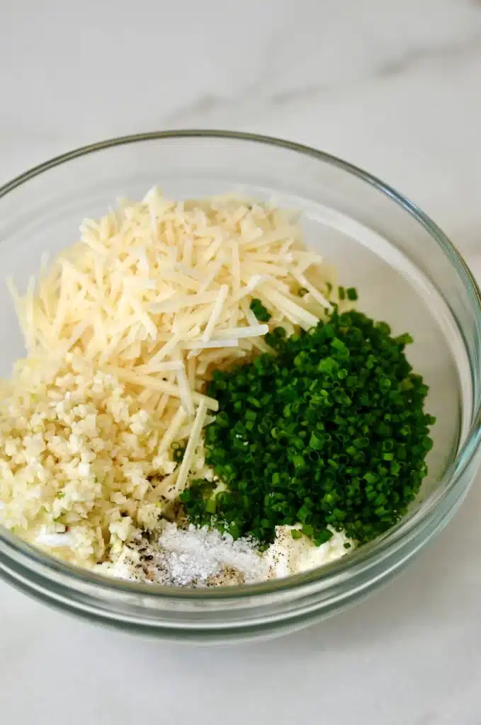 Glass bowl with grated cheese, minced garlic, chopped chives, and seasoning on a marble surface.