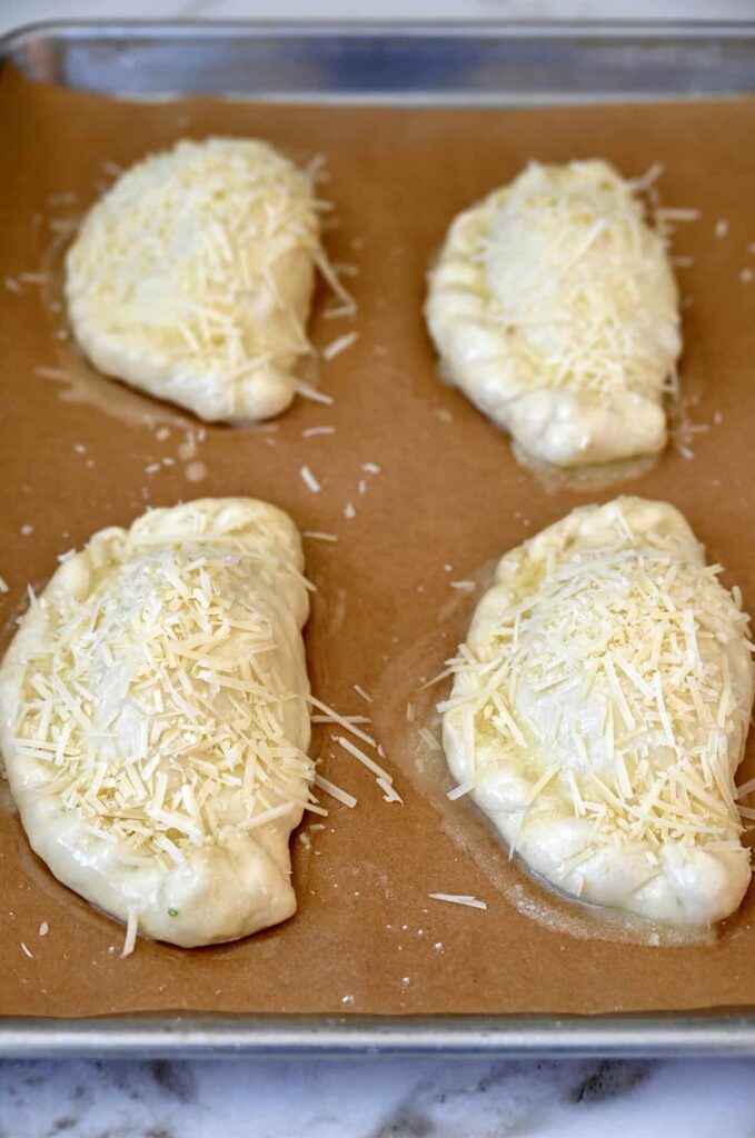 Unbaked calzones topped with shredded cheese on parchment paper, ready for the oven.