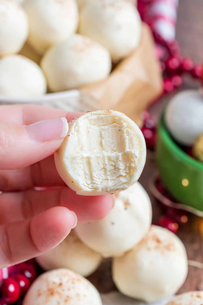 Close-up of a hand holding a creamy white chocolate truffle with a bite taken out, festive background.