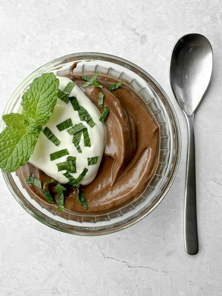 Chocolate mousse topped with cream, mint leaves, and shaved chocolate in a glass bowl beside a spoon on a marble surface.
