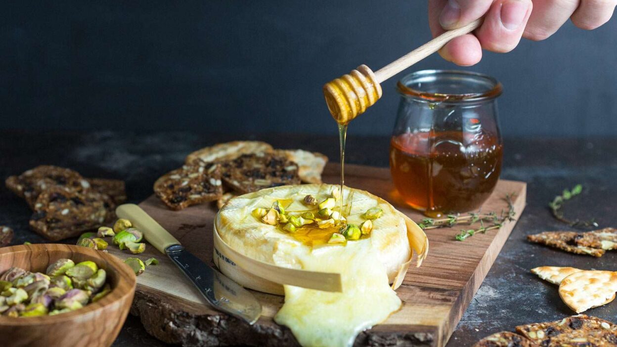 __opt__aboutcom__coeus__resources__content_migration__serious_eats__seriouseatscom__recipes__images__2017__01__20170124-Baked-Brie-honey-pistachios-1-matt-emily-clifton-0c2f407f976a4f2485ac3d3a5e14a53e - Melted Brie with Honey & Pistachios in 20 Minutes Melting cheese with honey drizzle, pistachios, and crackers on a wooden board, perfect for a gourmet appetizer.