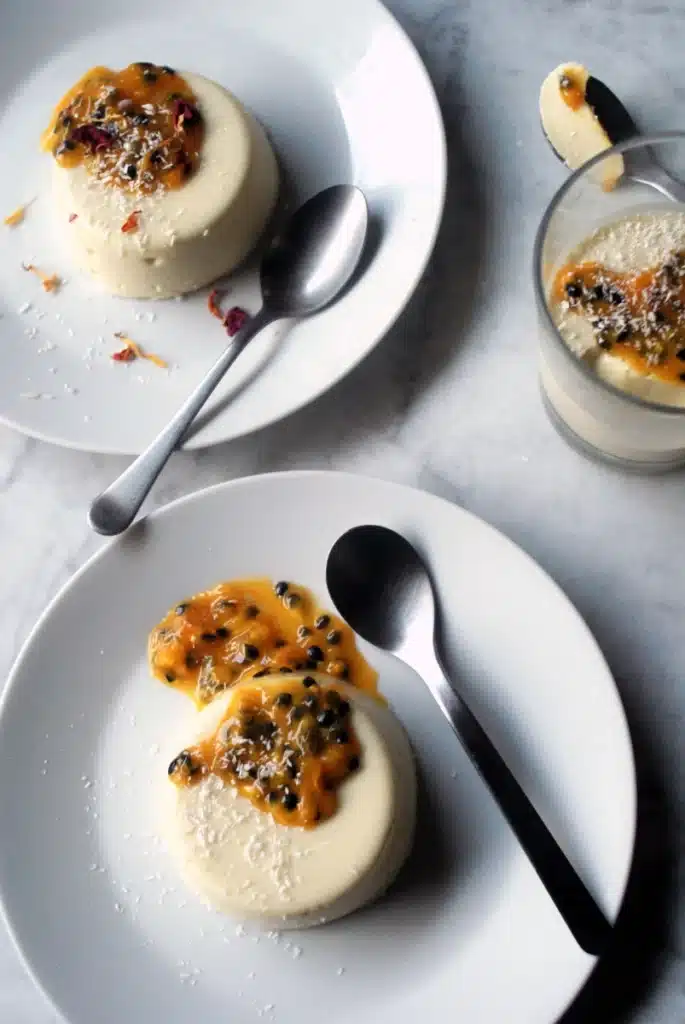 panna-cotta-x4-1 - Silky, Tropical Coconut & Passion Fruit Panna Cotta Delicious panna cotta topped with fresh passion fruit on white plates, served with spoons on a marble table.