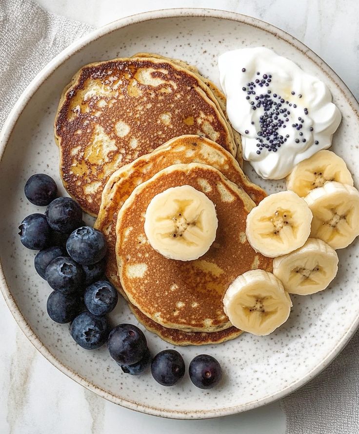 Fluffy pancakes with banana slices, blueberries, and yogurt on a ceramic plate for a delicious breakfast.