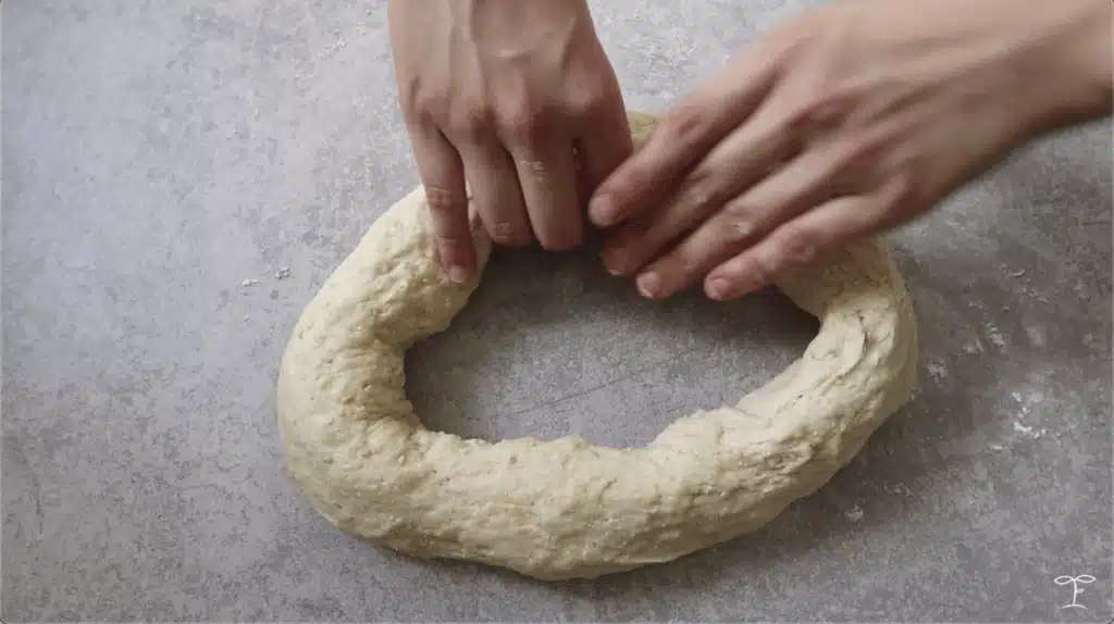 Hands shaping raw dough into a bagel on a floured surface.